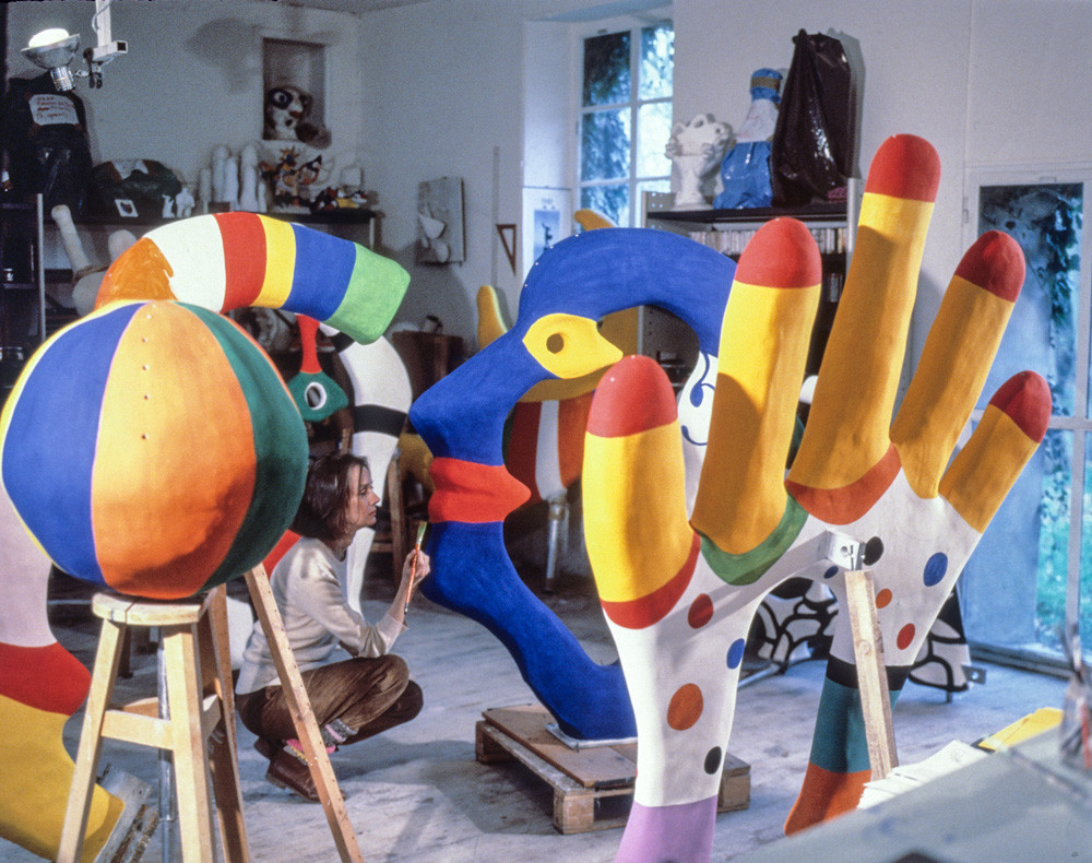 Artist Niki de Saint Phalle squads on the floor at her studio in Soisy surrounded by colorful sculptures. She holds a brush in her right hand aiming to paint an element sculpture tiled "Profile" for "Fontaine Chateau-Chinon", a collaborative project with Jean Tinguely, located in France.