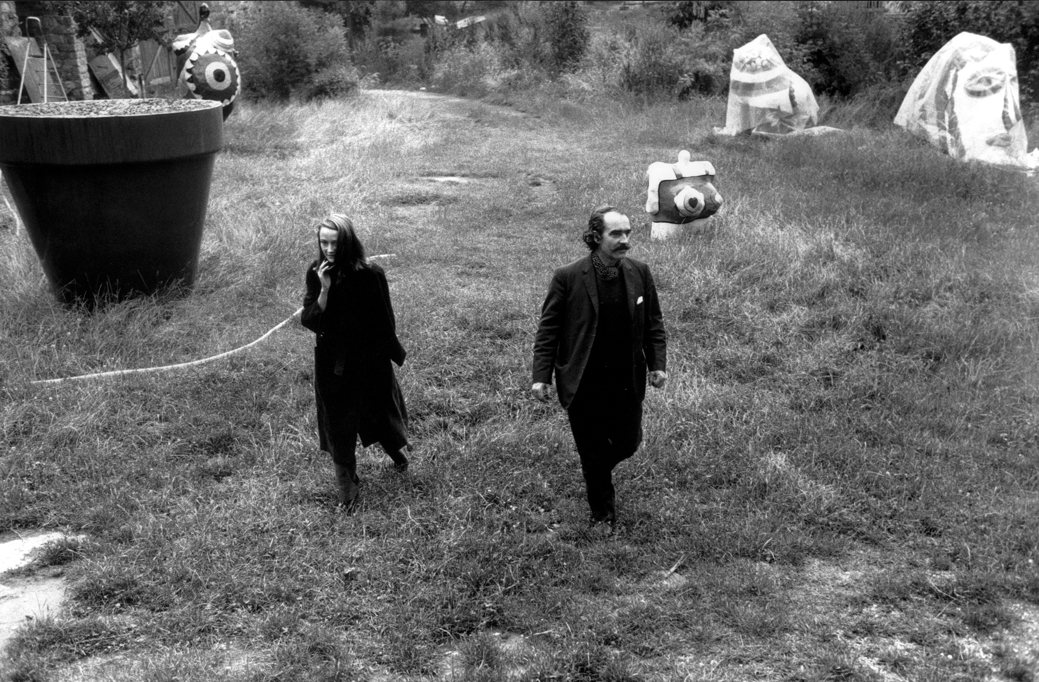 Garden setting with Saint Phalle's sculptures in the background. In the forefront Niki de Saint Phalle and Jean Tinguely walk toward the camera.